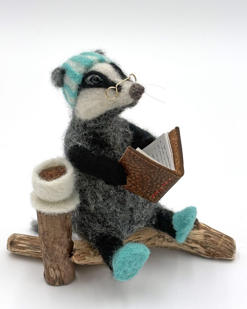 Needle felted anthropomorphic mixed media badger sculpture.  Figure wearing striped nightcap, glasses and slippers reads a book, and has a cup of tea. natural wood base