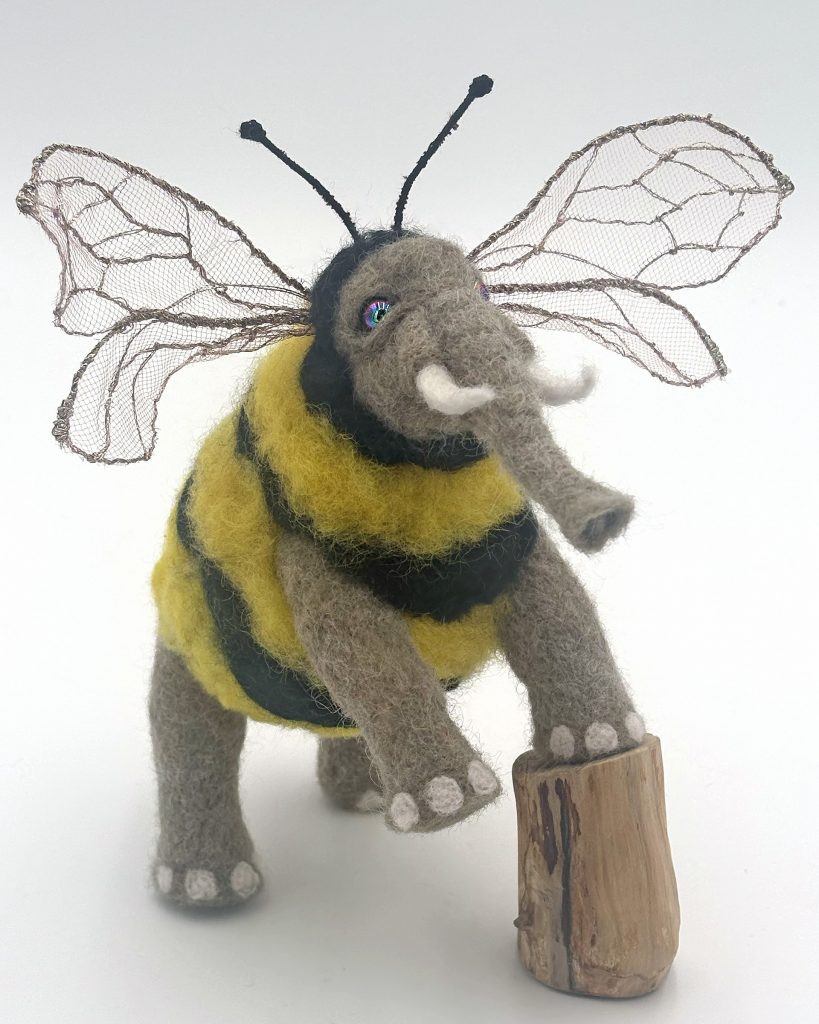 Needle felted bee-elephant chimera sculpture by Lynn Wartski . Grey elephant parts with yellow and black striped bee torso and gold toned embroidered tulle wing/ears.  Stands up on a natural wood stump