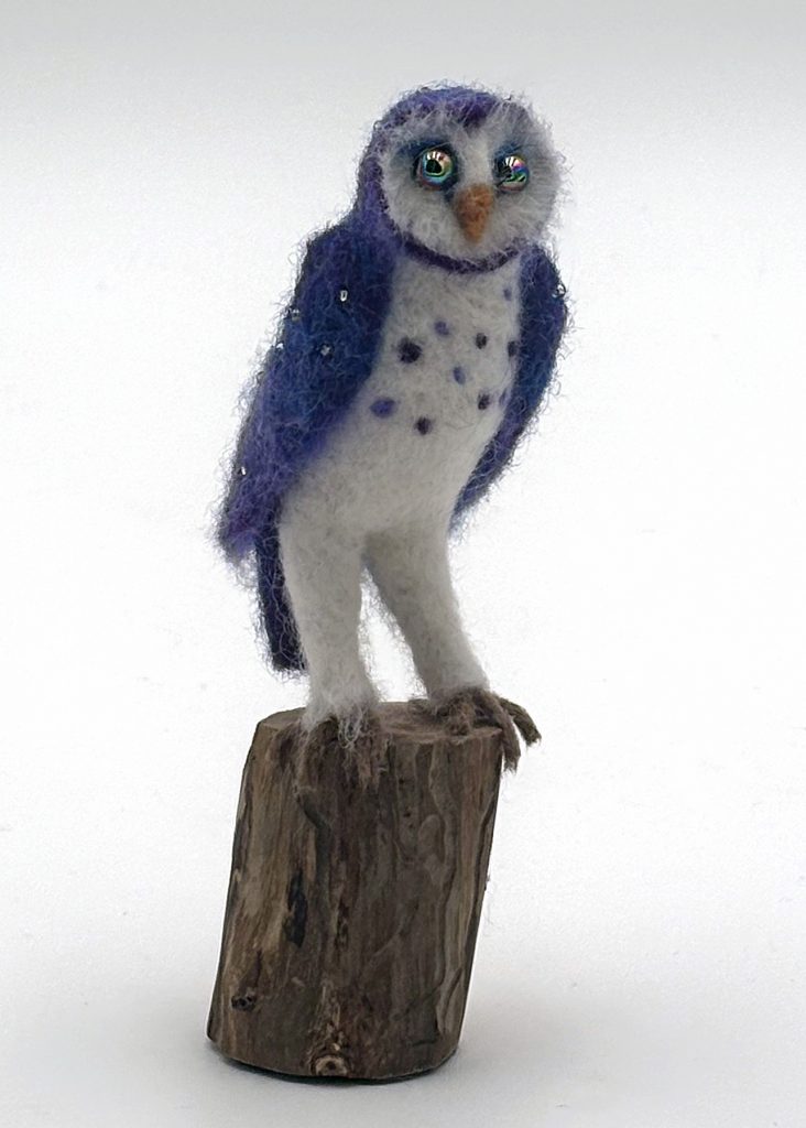 Anthropomorphic needle felted purple own sculpture by Lynn Wartski. Purple and midnight blue  owl with white chest, legs and face.  Glass bead eyes, and crystals sewn on back and upper wings. Stands on a natural wood base