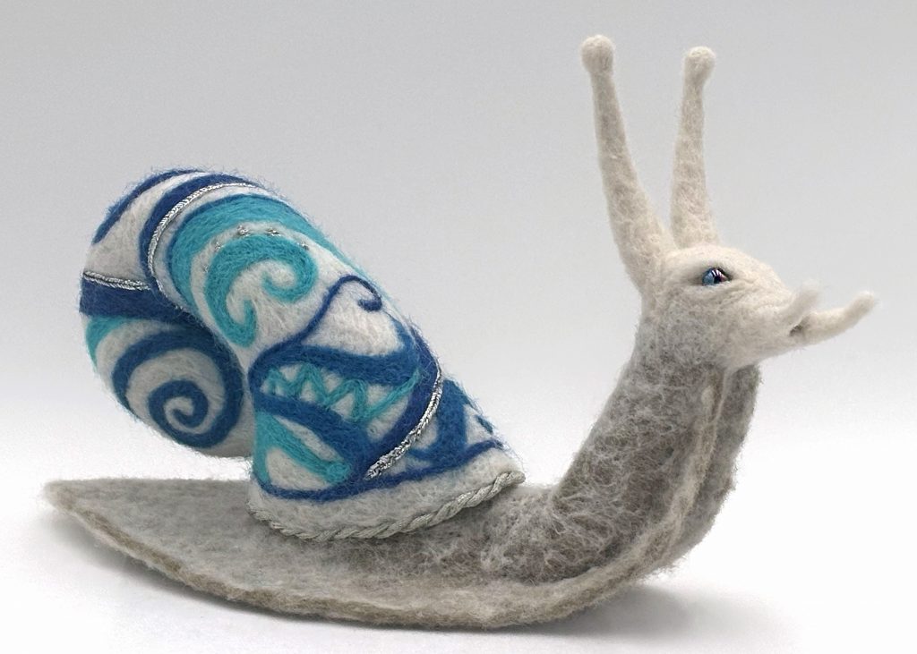 Needle felted snail sculpture by Lynn Wartski . Grey and green twith natural overlay snail body.  Shell is white with swirling and geometric pattern in shades of blue with silver metalic trim and embroidery