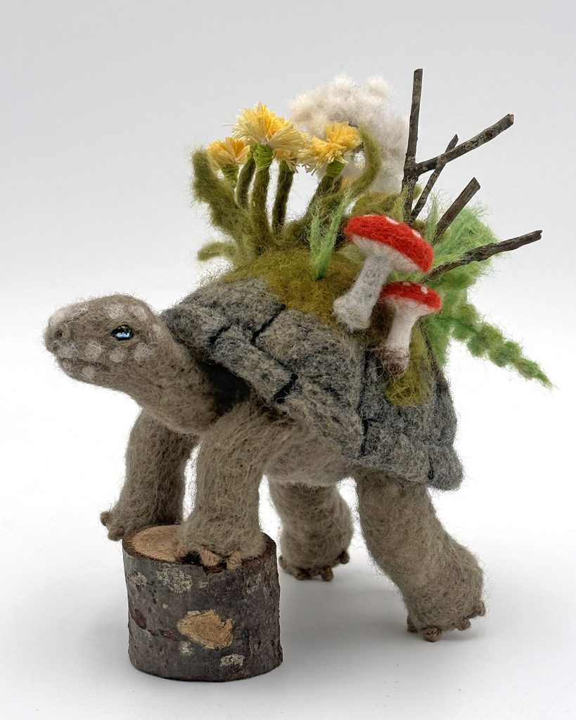 Needle felted tortoise with woodland flora sprouting from back.  A nod to turtle creation myths from around the world.  Grey and green tortoise figure with dandelion, ferns, moss branches and mushrooms in greens, yellow, white and red.