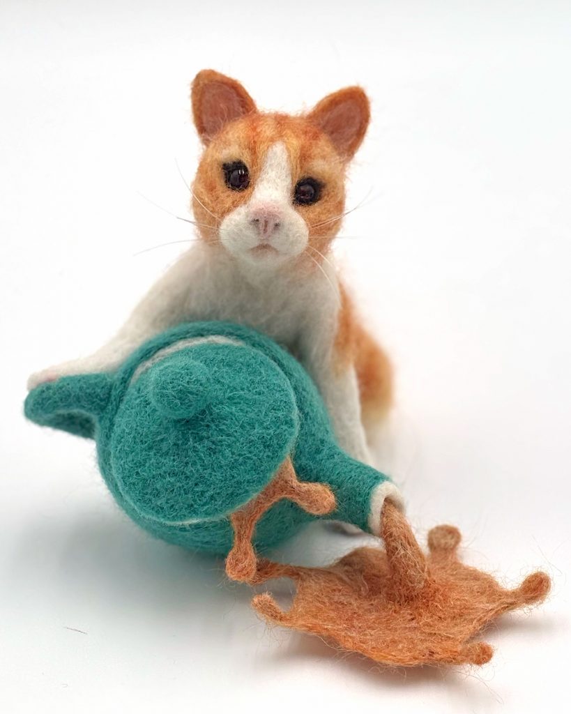 Anthropomorphic needle felted cat spilling a teapot sculpture by Lynn Wartski. Gold and white tabby kitten with glass bead eyes pushes over jade green teapot.  Felted "tea" captured mid spill comes out of spout and from under lid.