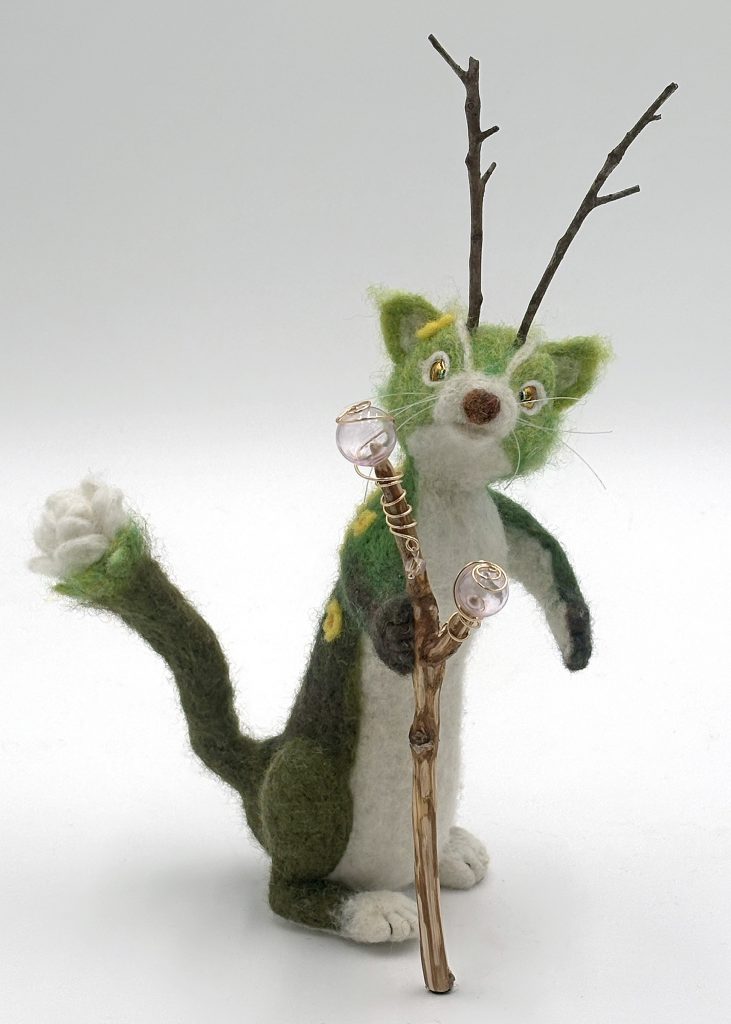 Bloom at Night needle felted anthropomorphic mythical creature sculpture by Lynn Wartski. Standing figure with gradation of green and a white belly and snout. Natural stick antennae and yellow flower embellishments. Figure has a white blossom at the end of tail, and holds a natural wood staff with blown glass and jewelry wire. elements