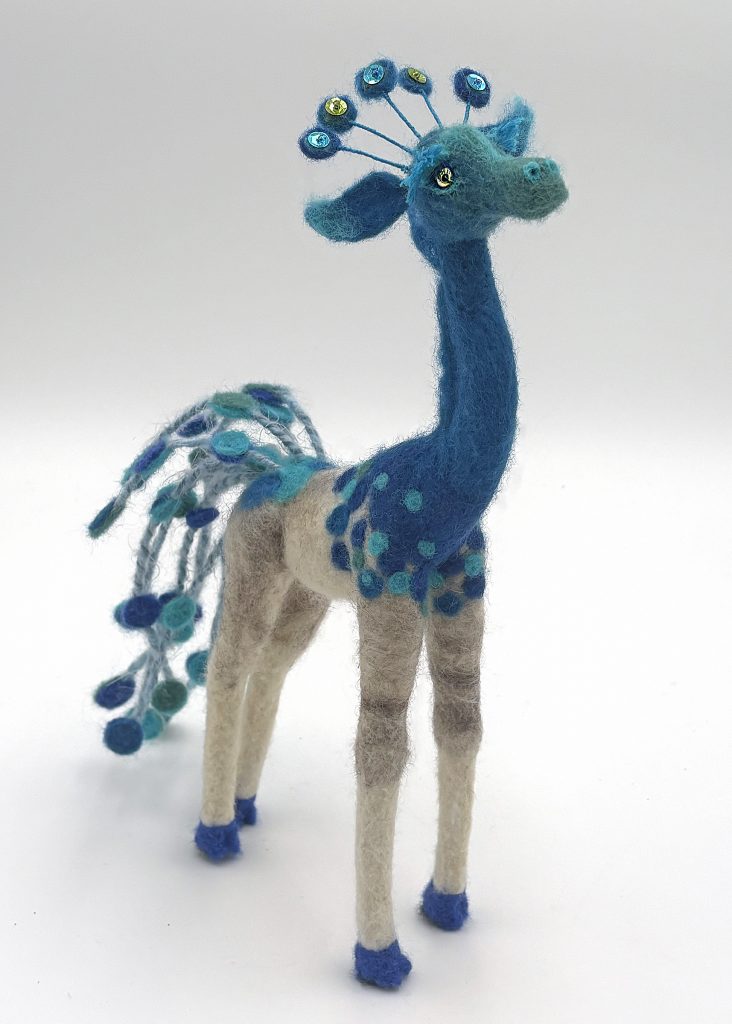 Qilin needle felted mythical animal figure sculpture by Lynn Wartski. inspired by creature from Chinese mythology. This form features a giraffe like animal with peacock color and patterning up the head and neck, and on its tail.
