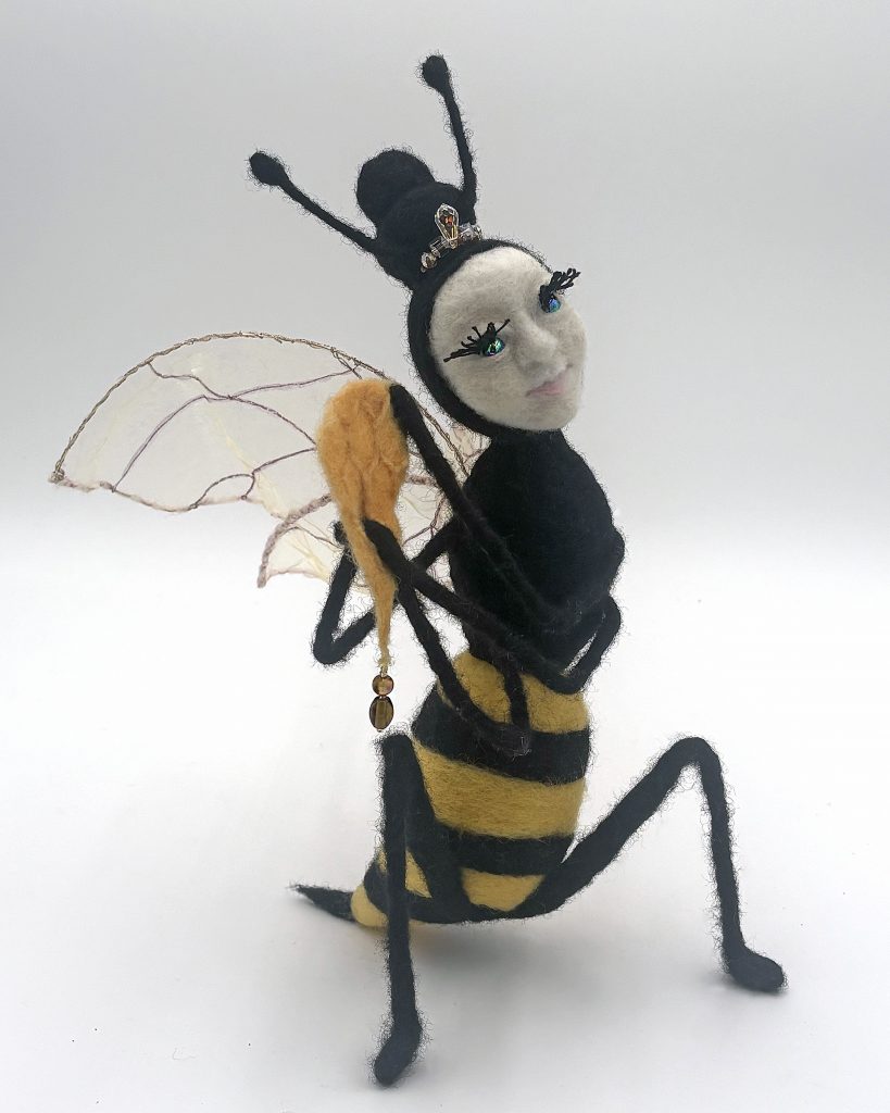 Honey needle felted figure sculpture by Lynn Wartski. Seated figure combines bee coloring (black and yellow) and form with a human face. Features embroidered tulle wings, and a crystal bead tiara infront of "bee hive" hair. Iridecent glass bead eyes, and holds a bit of honecomb with glass bead hone drip.