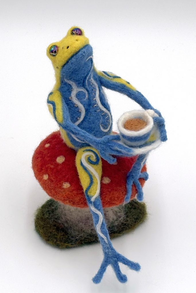 Tea needle felted anthropomorphic frog figure sculpture by Lynn Wartski. Seated figure in blue and yellow with white, dark blue felted and metalic embroidery embellishment. Sits on a red topped toadstool with gold dots, and holds a blue, yellow and white cup of tea on one knee