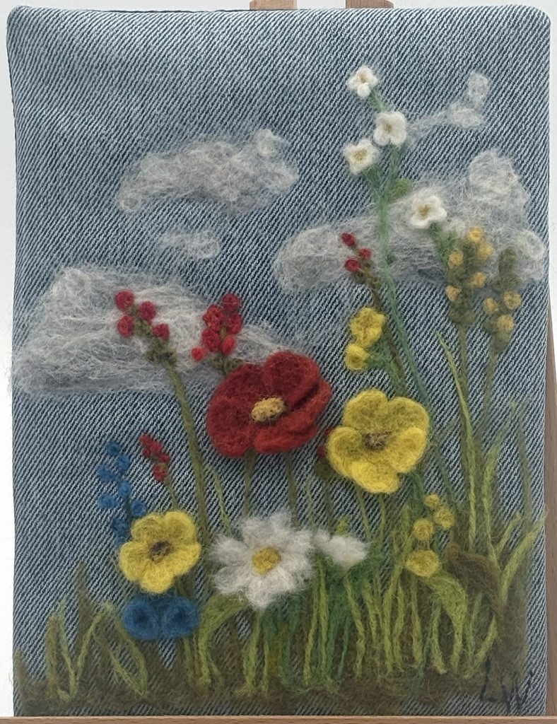 Needle felted wild flower meadow landscape on recycled denim.  red, white, yellow amd blue wildflowers with milti green foliage and white fair weather clouds.