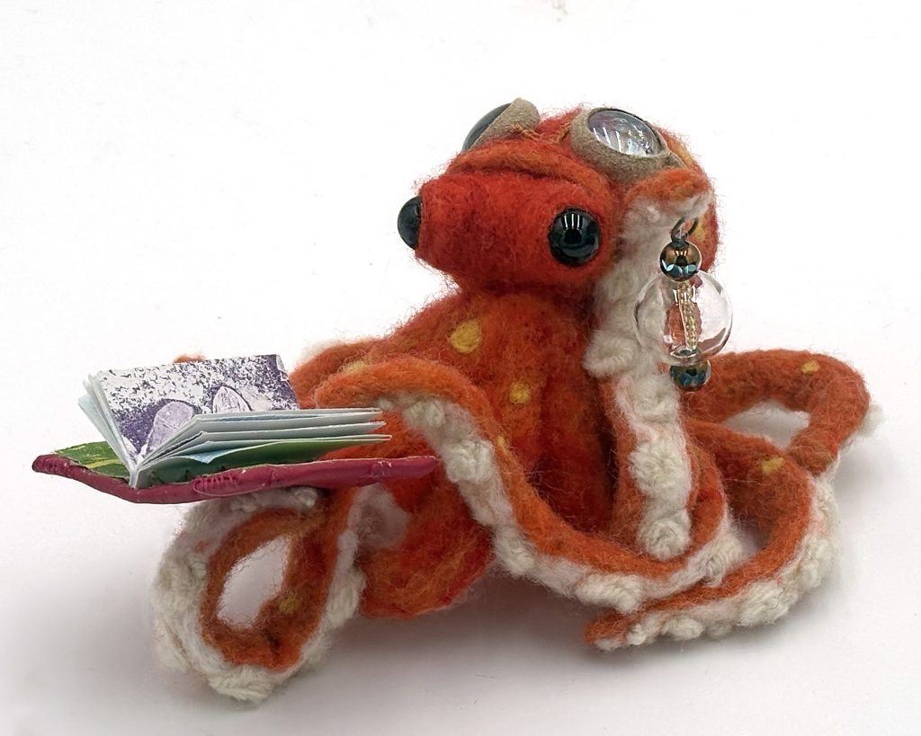 Remarkably Bright (2026) one-of-a-kind needle felted anthropomorphic steampunk octopus by Lynn Wartski.  Orange with yellow dots octopus figure holds a handmade book with red fux leather cover and botanical monoprint pages, and a lantern from glass and blown glass beads. Goggles fashioned from glass gems, upcycled suede, and jewelry findings.