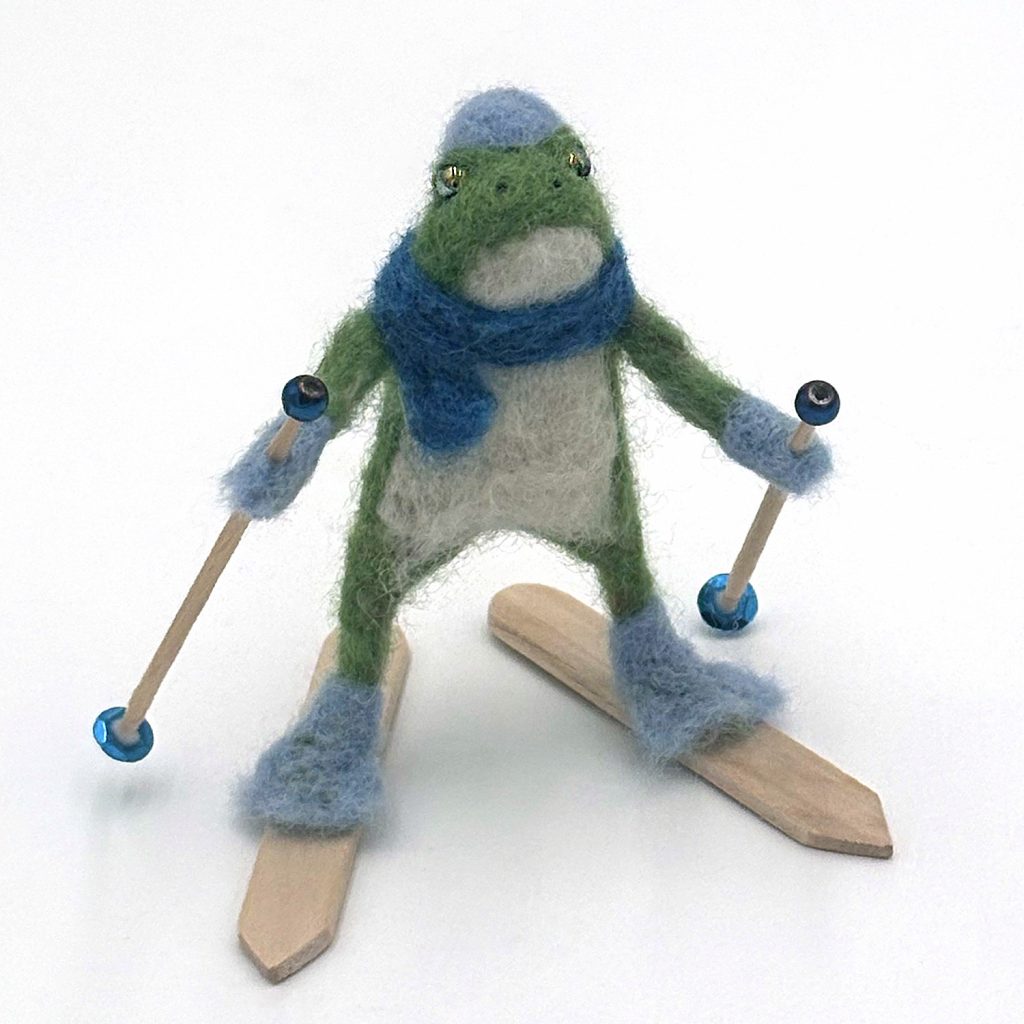 needle felted one-of-a-kind holiday ornament by Lynn Wartski. Frog on craft stick skis with toothpick poles, blue hat scarf, mittens and boots